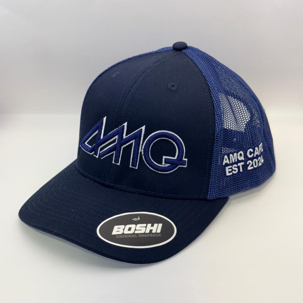 A navy blue trucker hat with a structured front panel and mesh back. The front features a large stylized 'AMHQ' logo outlined in white, and the side displays 'AMHQ CAR ENGINES EST 2014' in white.